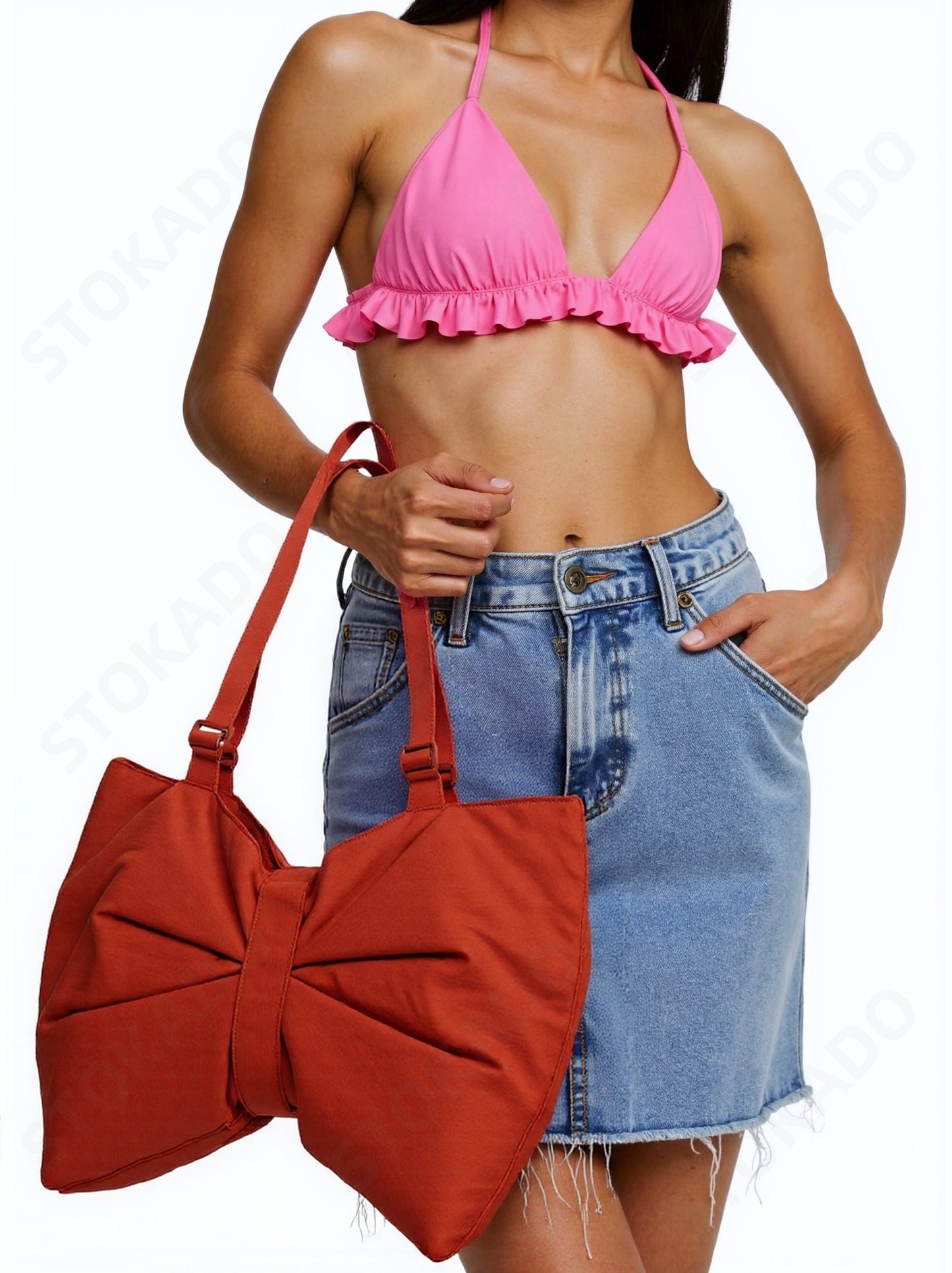 A woman wearing a pink ruffle crop top and denim skirt, carrying an oversized rust orange bow tote bag from STOKADO.